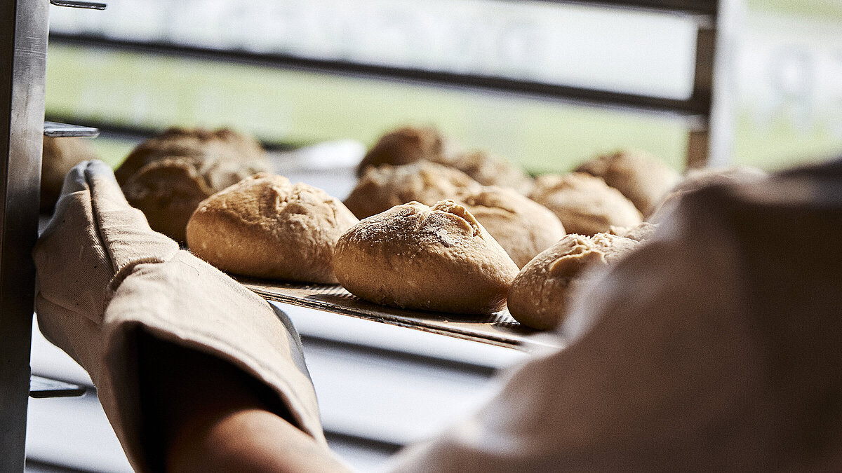 Blick über die linke Schulter eines Bäckers, der gerade Brötchen in eine Ablage schiebt