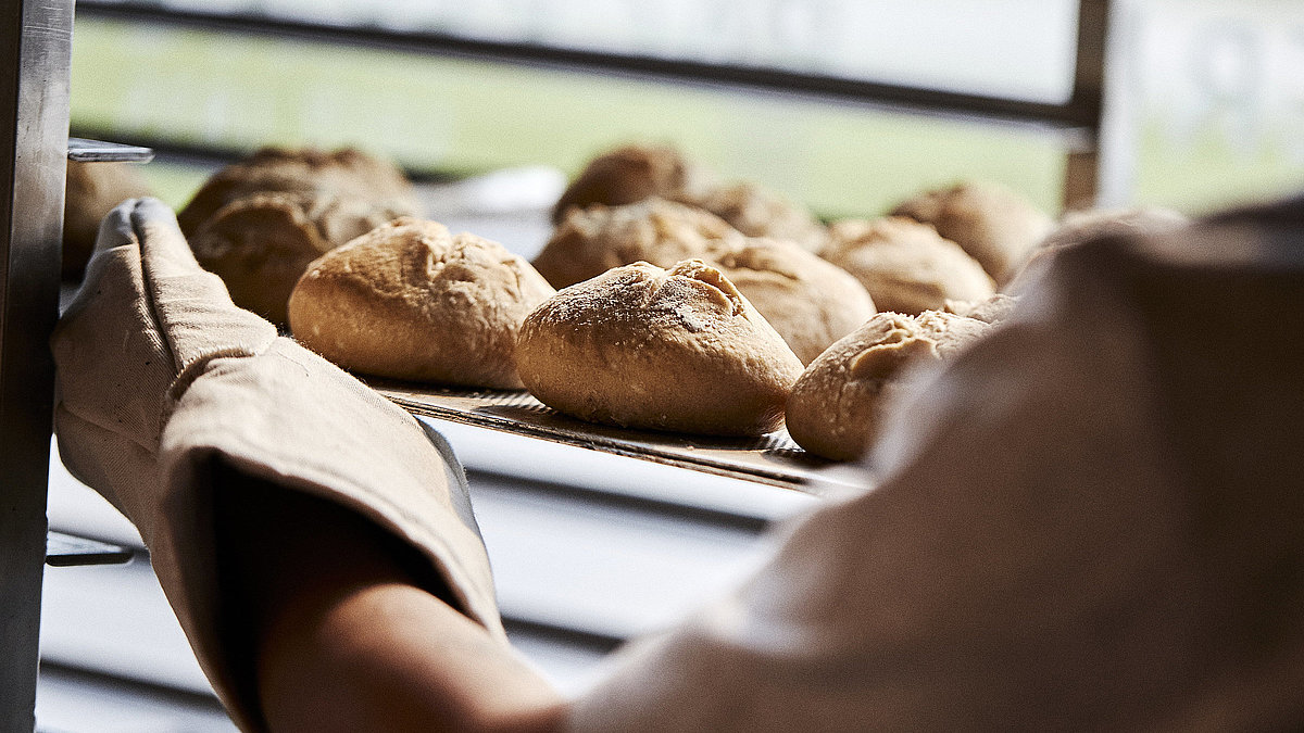 Blick über die linke Schulter eines Bäckers, der gerade Brötchen in eine Ablage schiebt