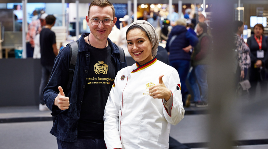 2 Personen auf der iba 2023.Links steht ein Mann, rechts eine Frau in Bäckerkleidung. Beide lachen in die Kamera und halten ihre Daumen nach oben