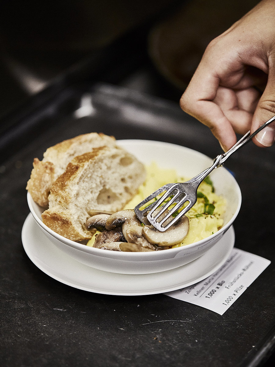 Person legt Champignons in einen Teller, in dem sich Rührei und zwei Scheiben weißes Brot befinden