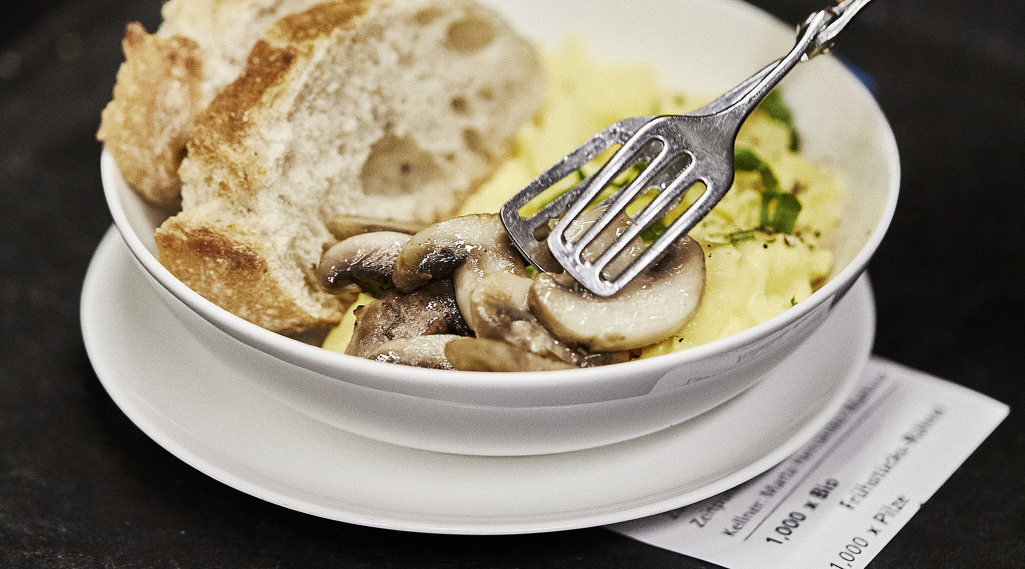Person legt Champignons in einen Teller, in dem sich Rührei und zwei Scheiben weißes Brot befinden