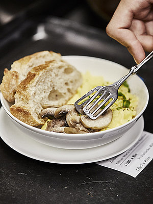 Person legt Champignons in einen Teller, in dem sich Rührei und zwei Scheiben weißes Brot befinden