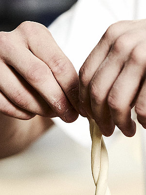 Bäcker formt eine Art Schleife aus Teig in der Luft. Jede Hand hält ein Teigende. 