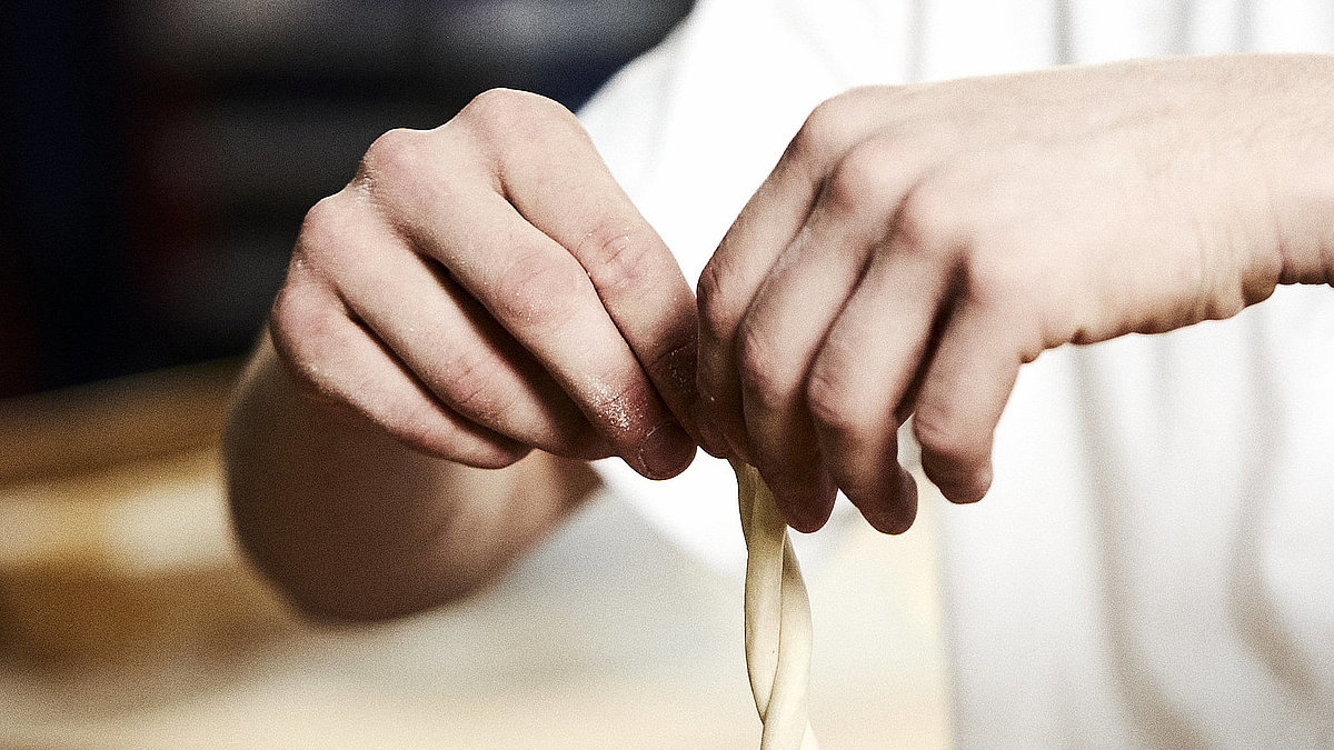 Bäcker formt eine Art Schleife aus Teig in der Luft. Jede Hand hält ein Teigende. 