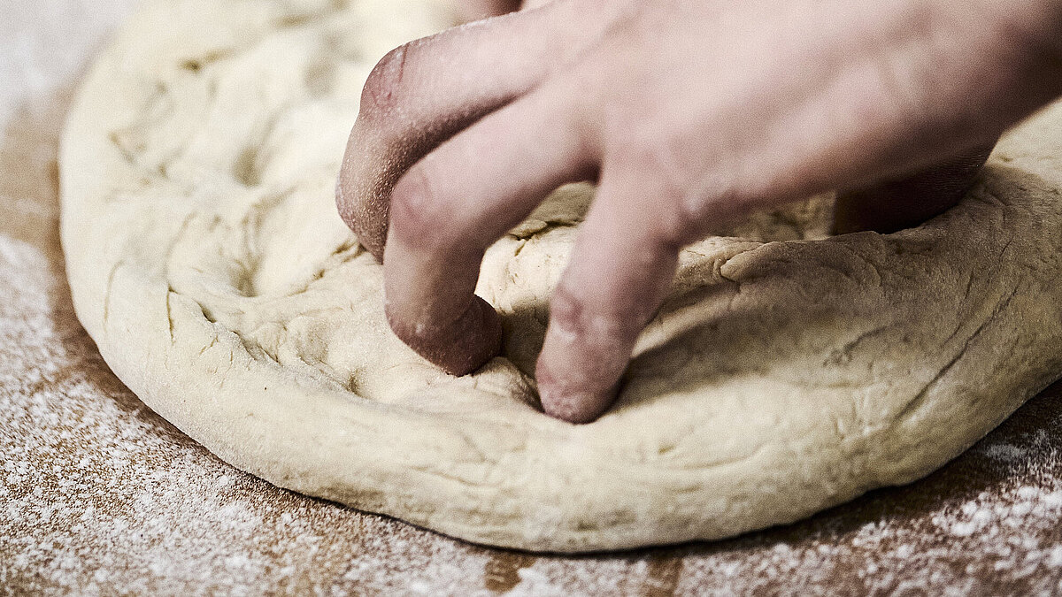 Bäckerhände bearbeiten einen flach ausgebreiteten Teig auf einer mehligen Arbeitsfläche. 