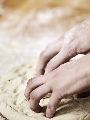 Bäckerhände bearbeiten einen flach ausgebreiteten Teig auf einer mehligen Arbeitsfläche. 