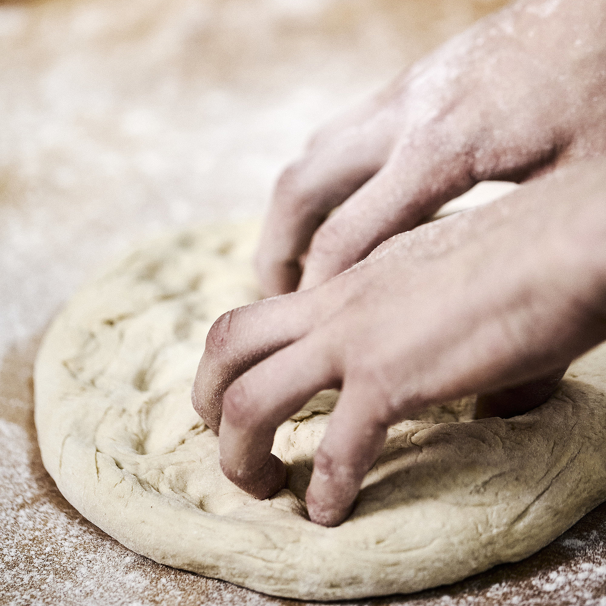 Bäckerhände bearbeiten einen flach ausgebreiteten Teig auf einer mehligen Arbeitsfläche. 
