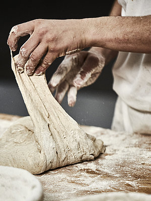 Bäckerhände verarbeiten einen Teig auf einer mehligen Arbeitsfläche. Im Vordergrund ist ein Korb zu sehen.