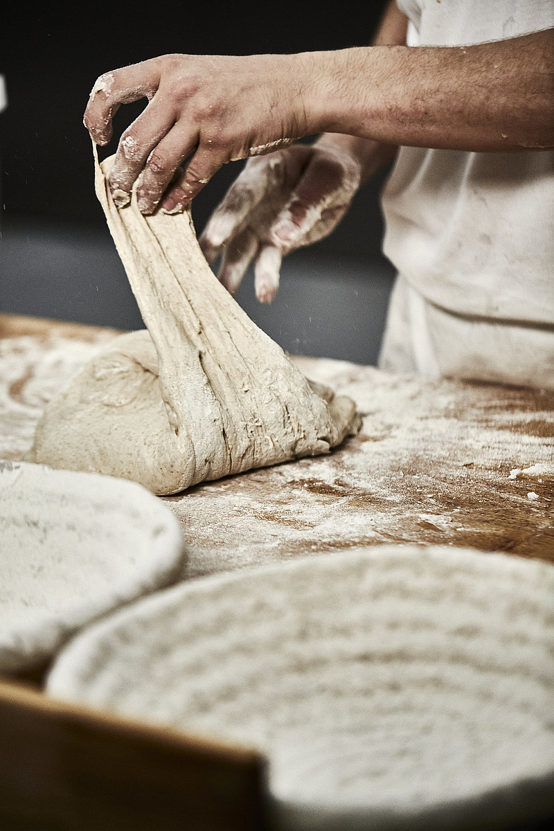 Bäckerhände verarbeiten einen Teig auf einer mehligen Arbeitsfläche. Im Vordergrund ist ein Korb zu sehen.
