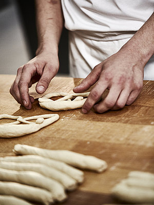 Bäckerhände formen eine Laugenbreze aus Teig. Im Vordergrund sind Teigrollen zu sehen.