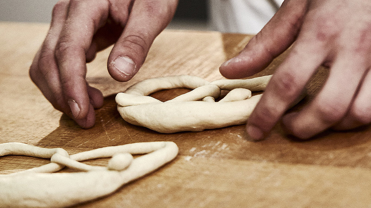 Bäckerhände formen eine Laugenbreze aus Teig. Im Vordergrund sind Teigrollen zu sehen.