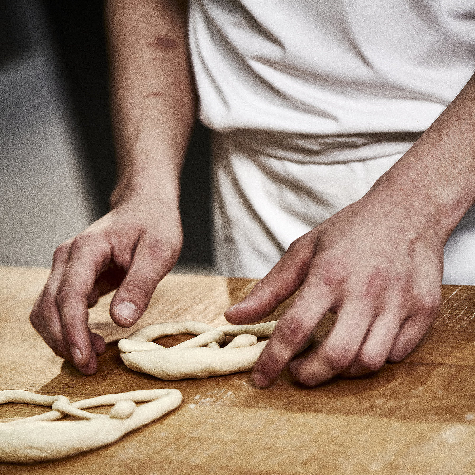 Bäckerhände formen eine Laugenbreze aus Teig. Im Vordergrund sind Teigrollen zu sehen.