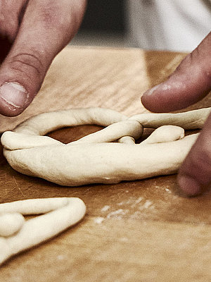 Bäckerhände formen eine Laugenbreze aus Teig. Im Vordergrund sind Teigrollen zu sehen.