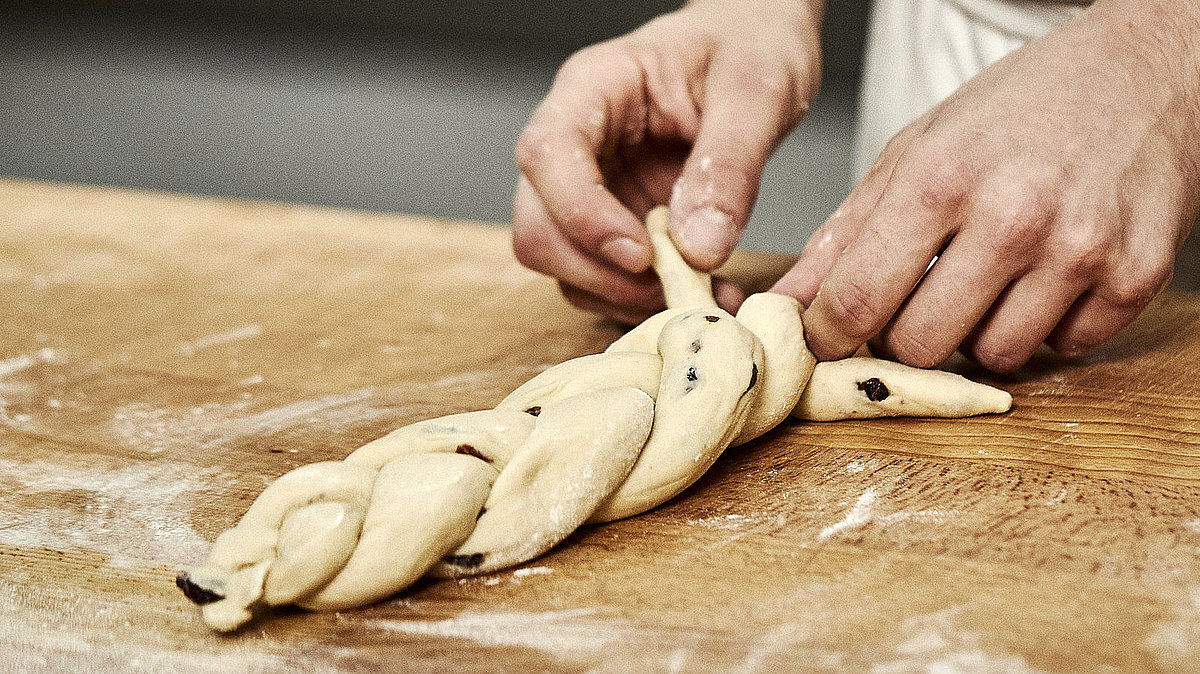 Bäckerhände flechten einen Zopf aus Teig auf einer mehligen Arbeitsfläche.