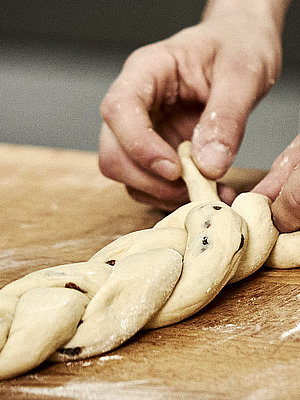 Bäckerhände flechten einen Zopf aus Teig auf einer mehligen Arbeitsfläche.