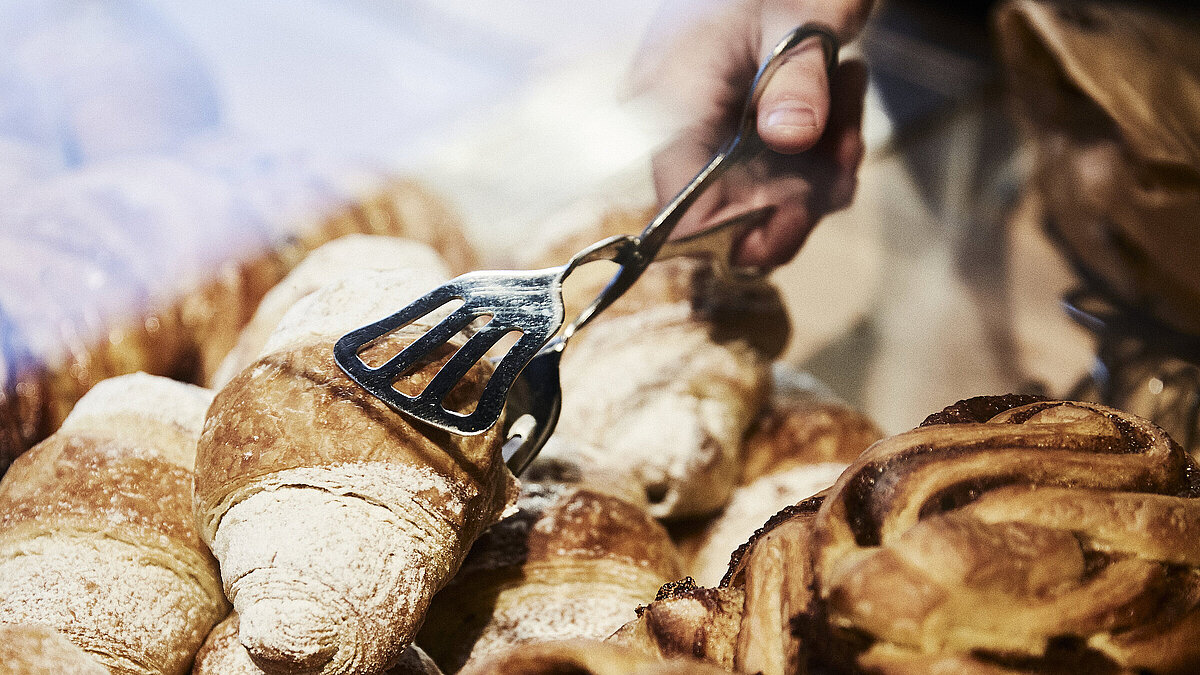 Person greift mit einer metallischen Gebäckzange nach eine Croissant, dass inmitten mehrerer süßen Backwaren liegt. 
