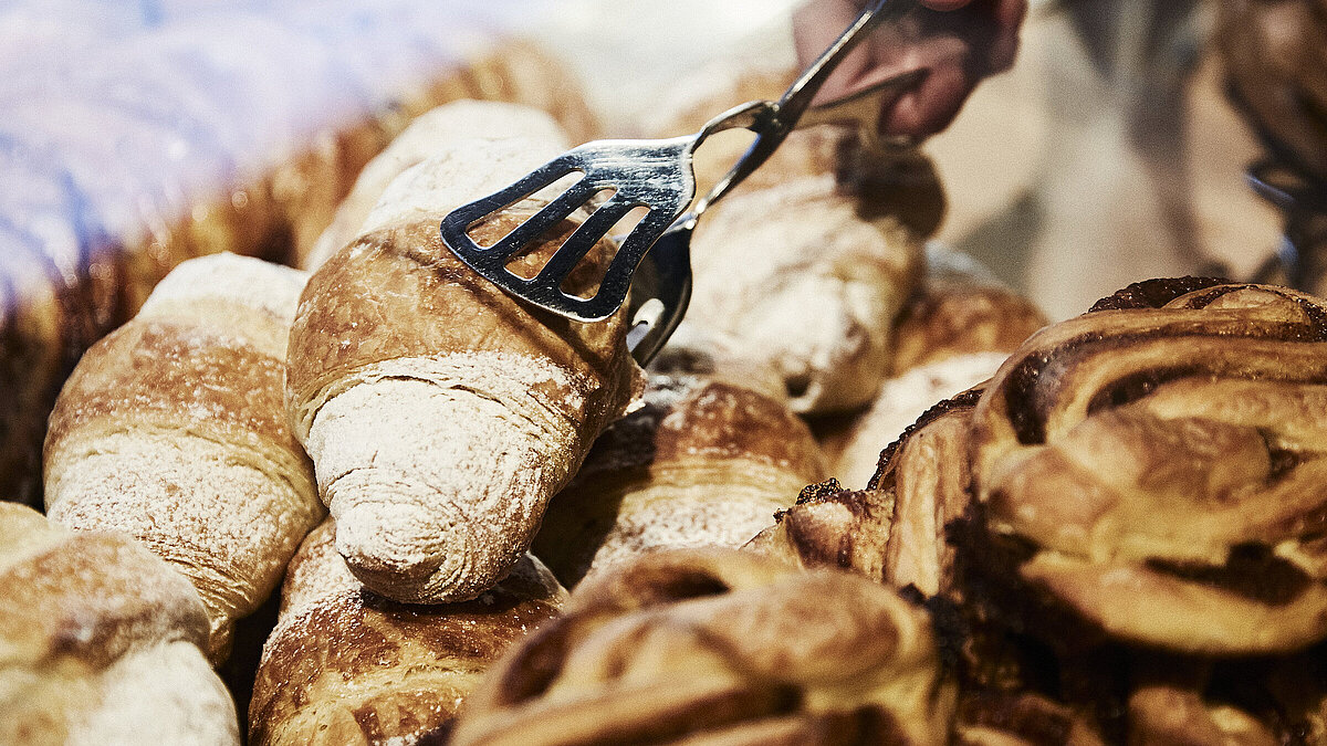 Person greift mit einer metallischen Gebäckzange nach eine Croissant, dass inmitten mehrerer süßen Backwaren liegt. 
