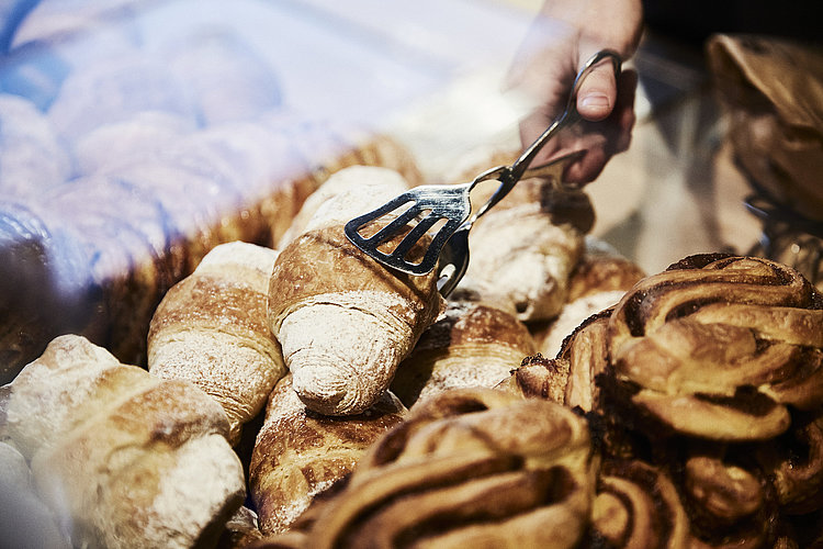 Person greift mit einer metallischen Gebäckzange nach eine Croissant, dass inmitten mehrerer süßen Backwaren liegt. 