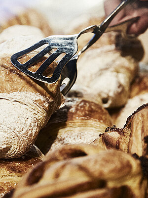 Person greift mit einer metallischen Gebäckzange nach eine Croissant, dass inmitten mehrerer süßen Backwaren liegt. 