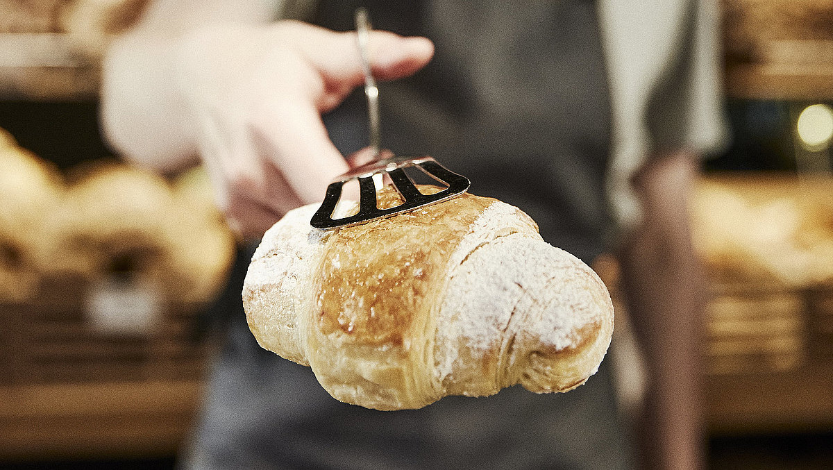 Weibliche Person mit grauer Kochschürze reicht ein Croissant in die Kamera, das Croissant ist scharf, die Person ist unscharf.