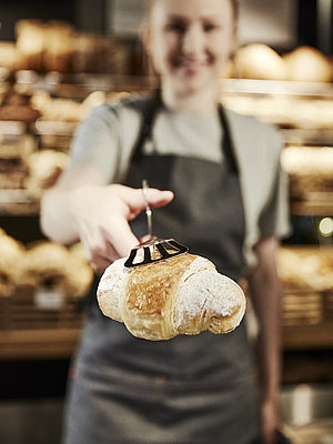 Weibliche Person mit grauer Kochschürze reicht ein Croissant in die Kamera, das Croissant ist scharf, die Person ist unscharf.