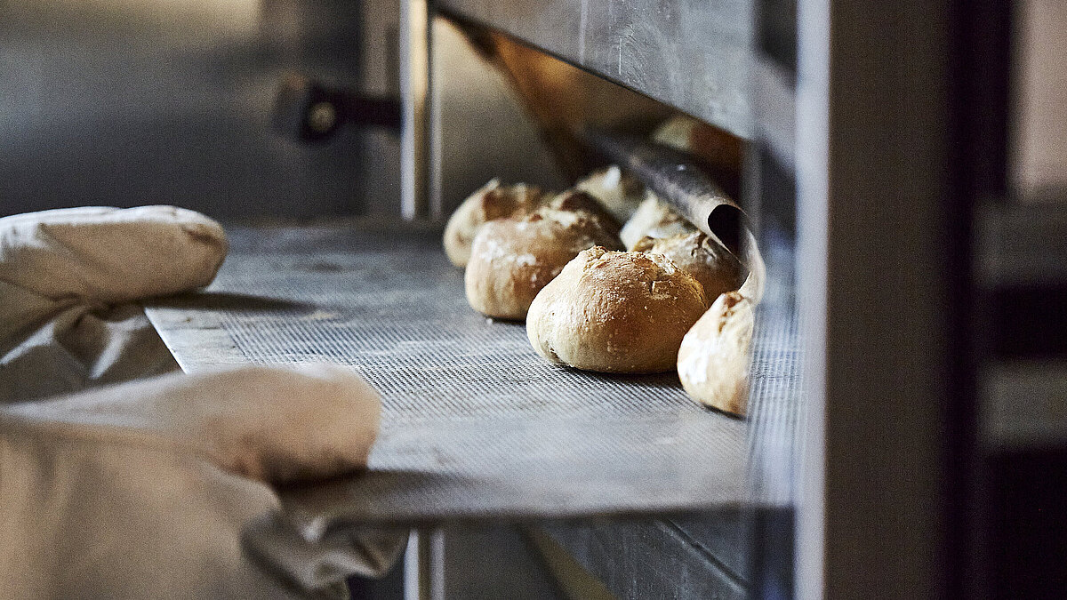 Bäcker mit großen Handschuhen schiebt ein Backblech mit Brötchen in den Ofen.