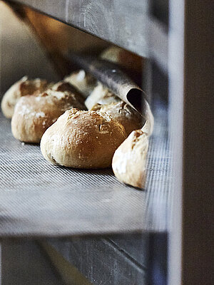 Bäcker mit großen Handschuhen schiebt ein Backblech mit Brötchen in den Ofen.
