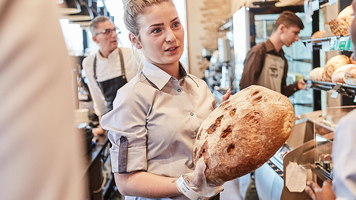 Bäckereifachverkäuferin spricht mit einer Kollegin und hält dabei ein Brot