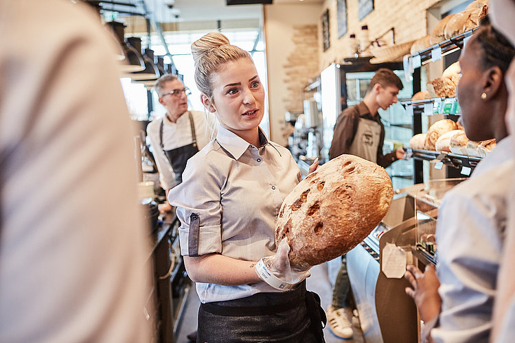 Bäckereifachverkäuferin spricht mit einer Kollegin und hält dabei ein Brot