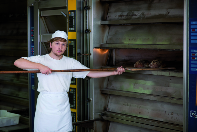 Bäckerauszubildender mit Broten auf Schießer am Ofen
