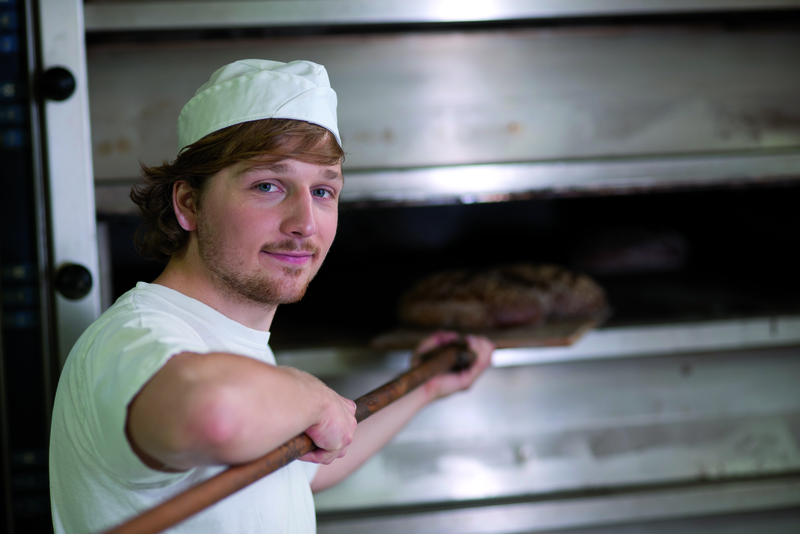 Bäckerauszubildender mit Brot auf Schießer am Ofen