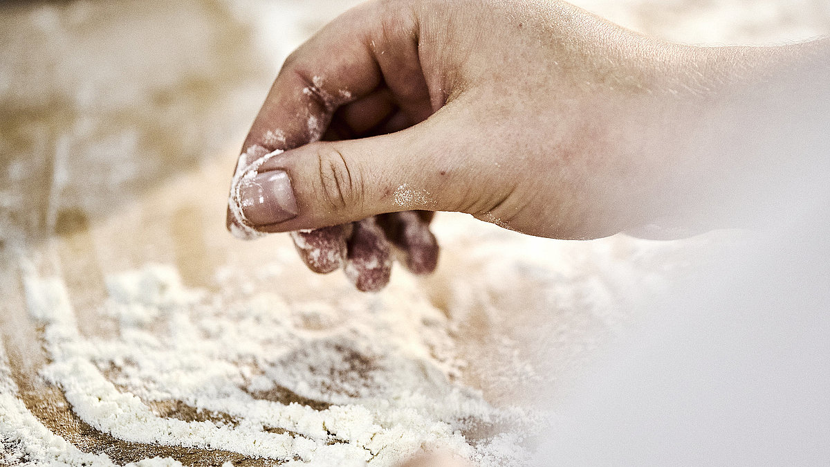 Detailaufnahme einer Bäckerhand, die Mehl auf einer Arbeitsfläche verteilt.