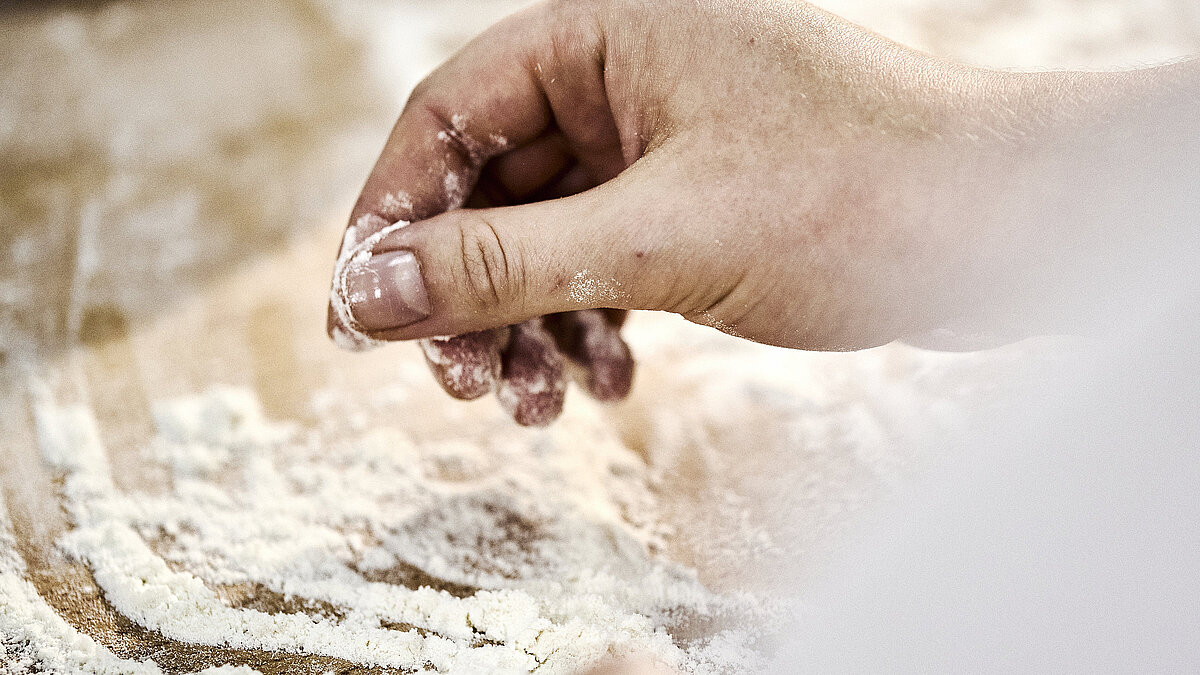 Detailaufnahme einer Bäckerhand, die Mehl auf einer Arbeitsfläche verteilt.